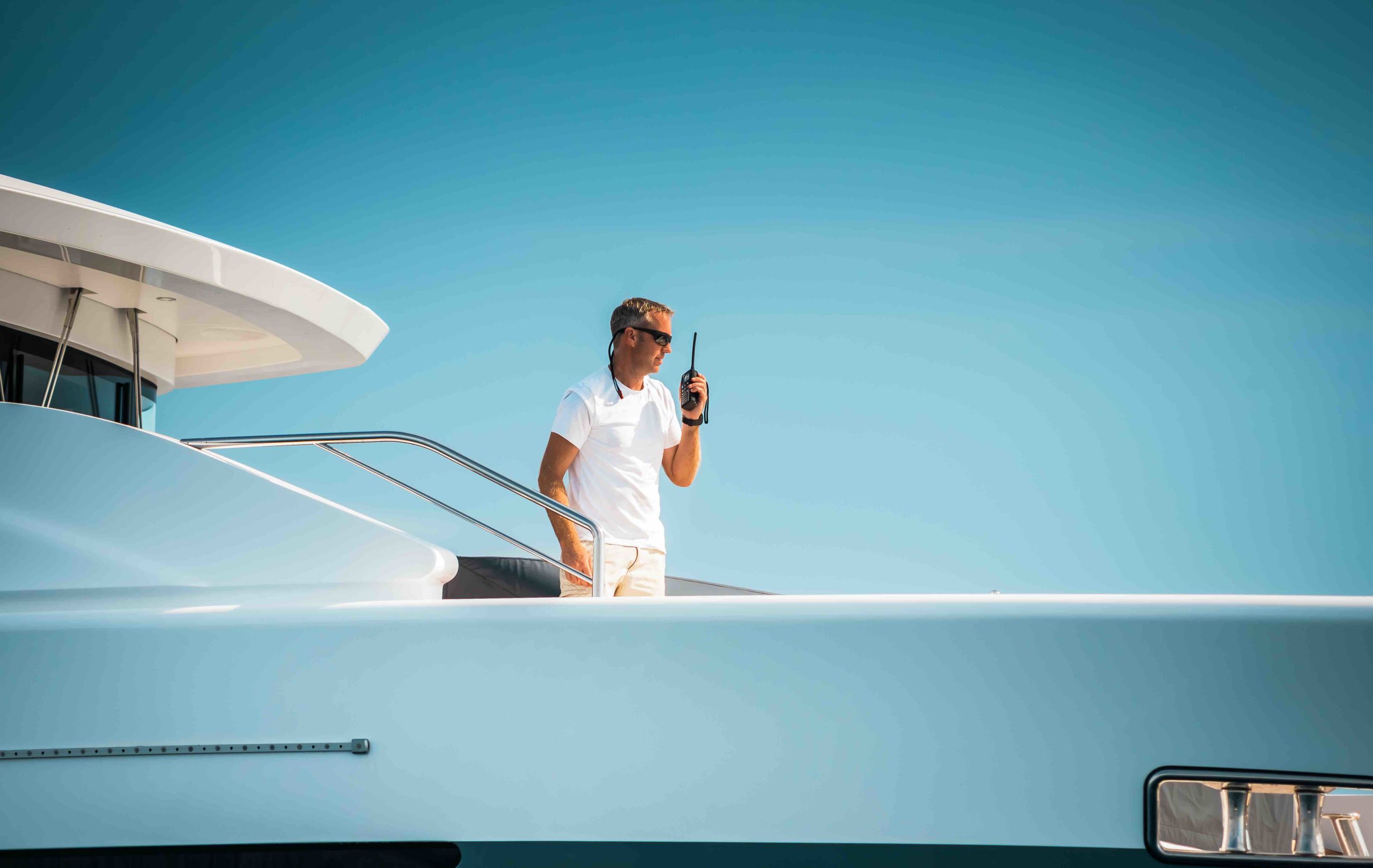 Male superyacht Deckhand with a handheld radio getting ready to drop anchor, with a blue sky in the background