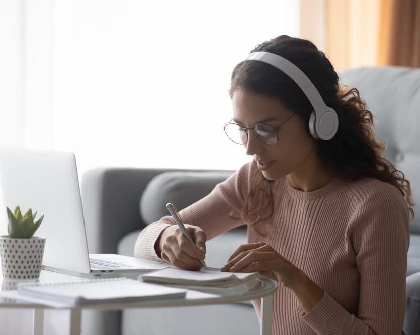 Focused woman wearing headphones writing notes, sitting on floor at home, serious girl female student wearing glasses learning language, listening to online course or music while doing homework