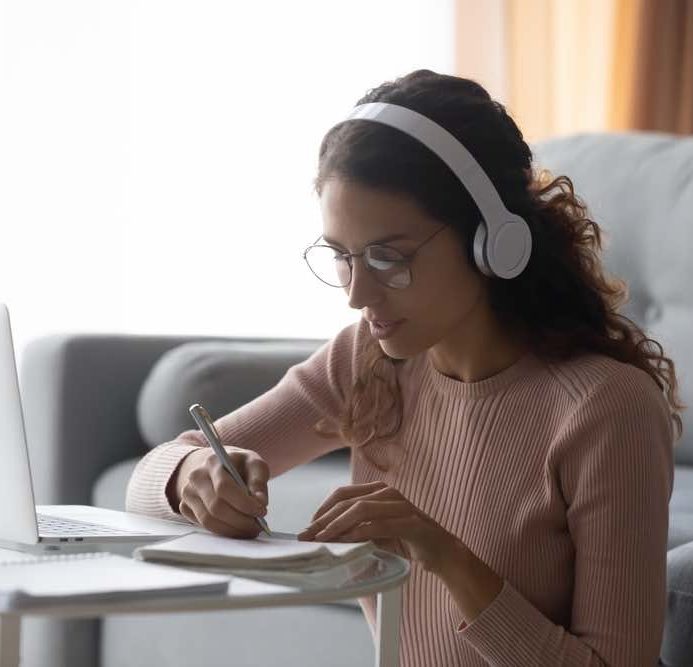 Focused woman wearing headphones writing notes, sitting on floor at home, serious girl female student wearing glasses learning language, listening to online course or music while doing homework