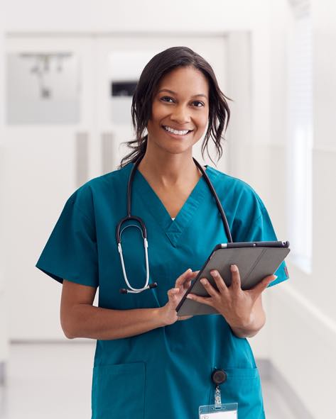 Portrait Of Smiling Female Doctor Wearing Scrubs In Hospital Corridor Holding Digital Tablet