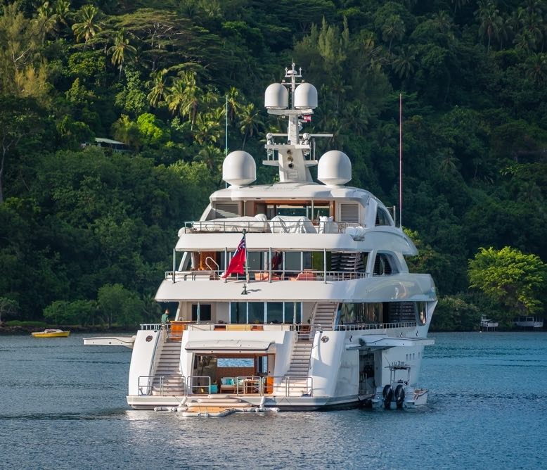 The stern of large sixty meter super yacht at anchor in Cook's Bay in tropical island of Moorea, French Polynesia. With verdant green landscape and large fishing tender in the water