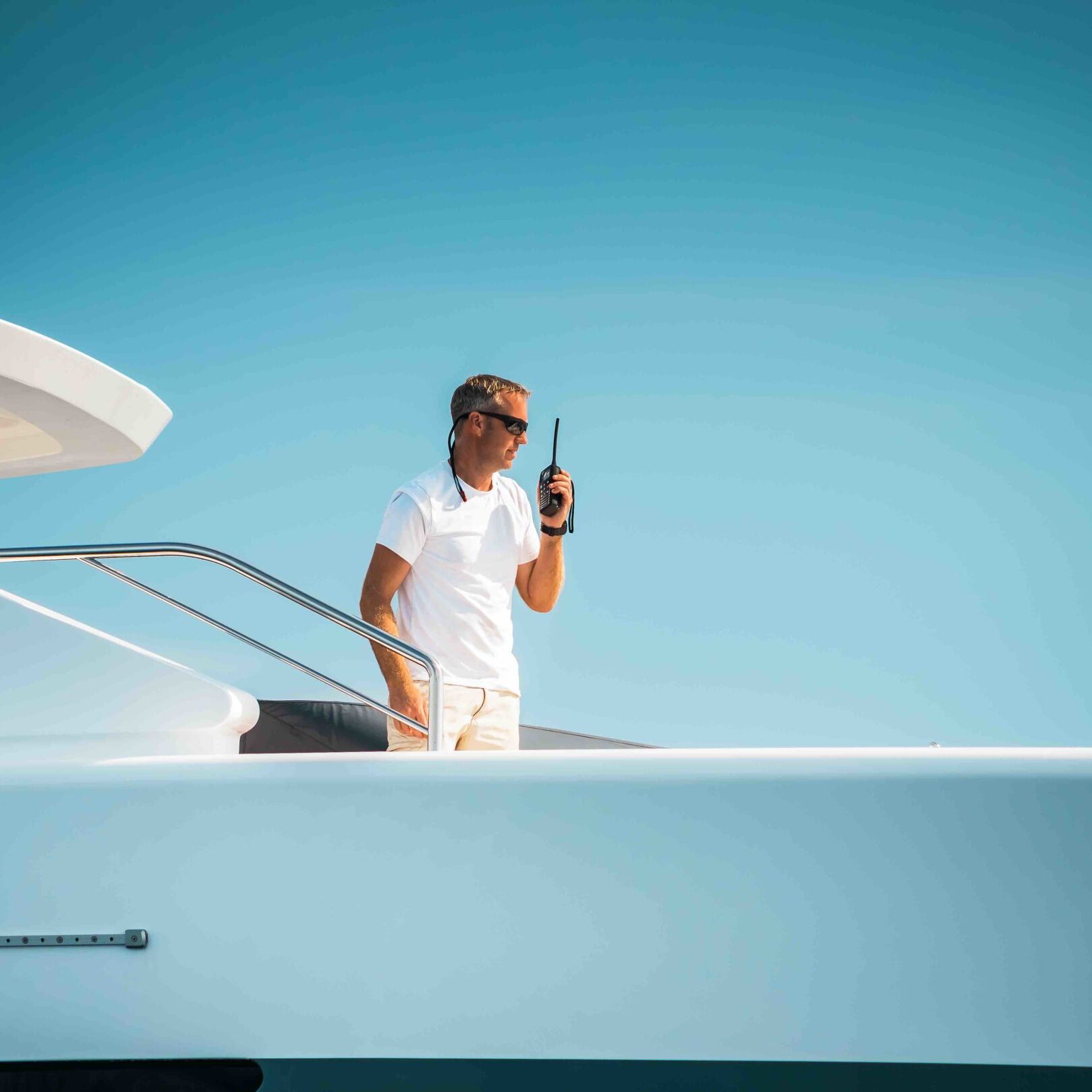 Male superyacht Deckhand with a handheld radio getting ready to drop anchor, with a blue sky in the background