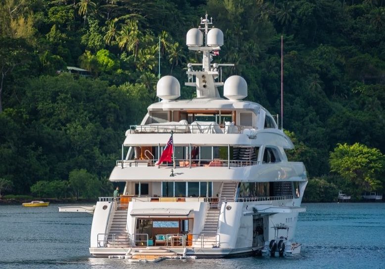 The stern of large sixty meter super yacht at anchor in Cook's Bay in tropical island of Moorea, French Polynesia. With verdant green landscape and large fishing tender in the water
