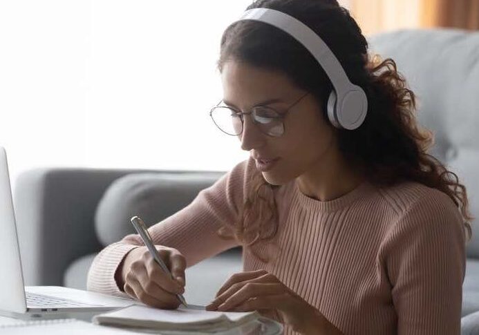 Focused woman wearing headphones writing notes, sitting on floor at home, serious girl female student wearing glasses learning language, listening to online course or music while doing homework